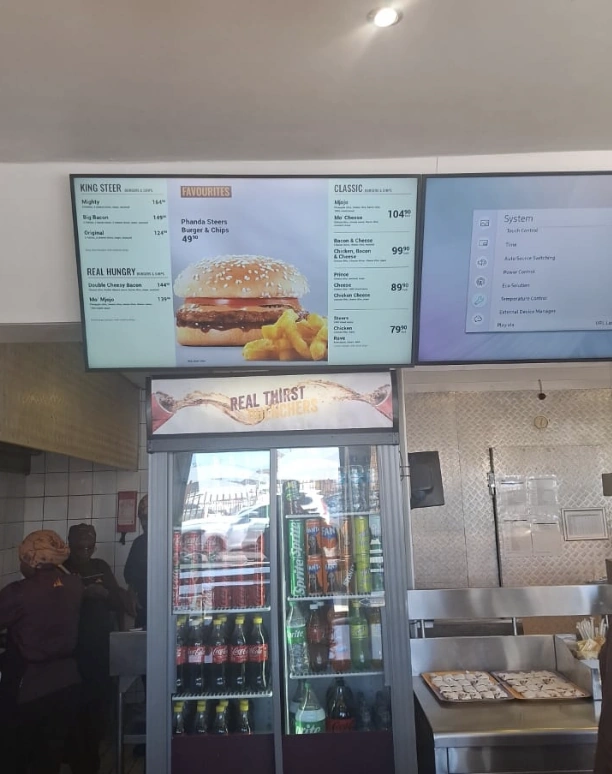 Fast-food restaurant with digital burger menu boards, glass drinks refrigerator, and stainless-steel preparation area.