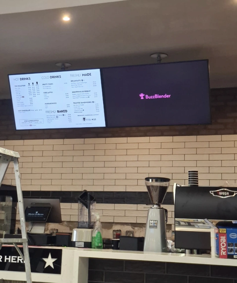 Cafe counter with Order Here and Collect Here signs, digital menu screens, food display cases, espresso machine, and staff.