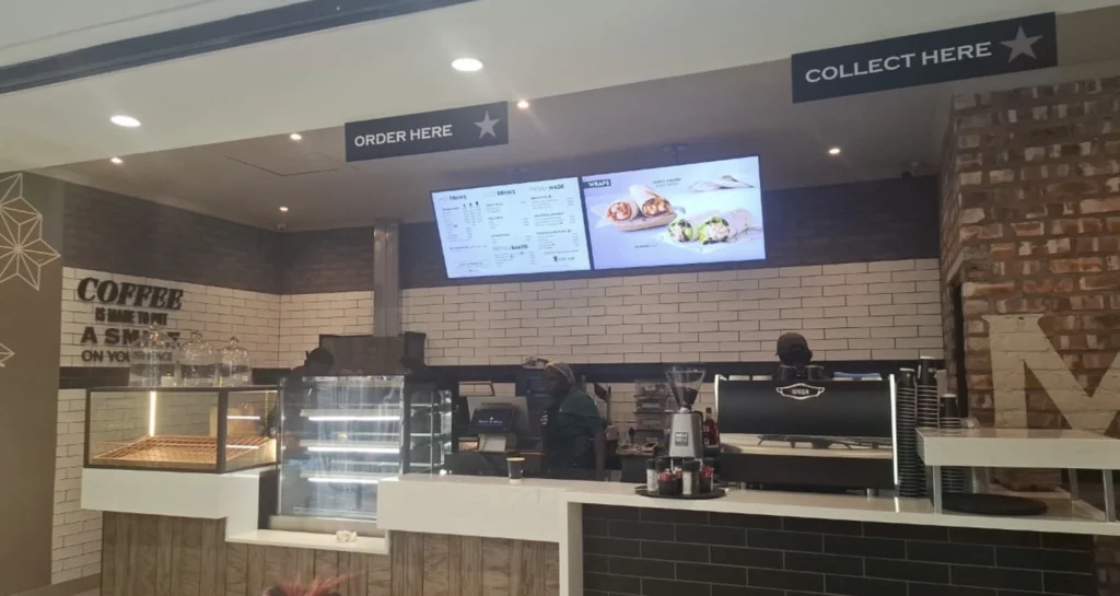 Coffee shop counter with two digital menu screens, espresso machine, coffee grinder, blender, and tiled wall.